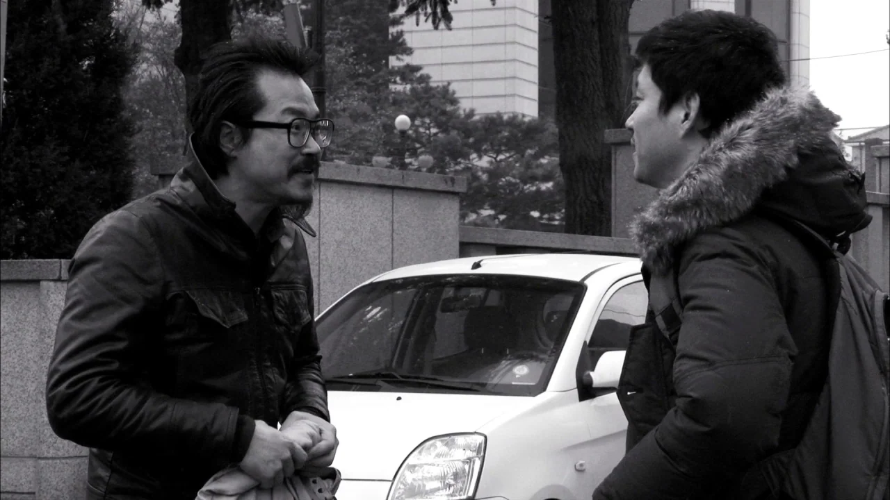 The Day He Arrives (2018) movie still: medium — An outdoor parking lot with concrete walls and a parked white car. Two men converse while standing…