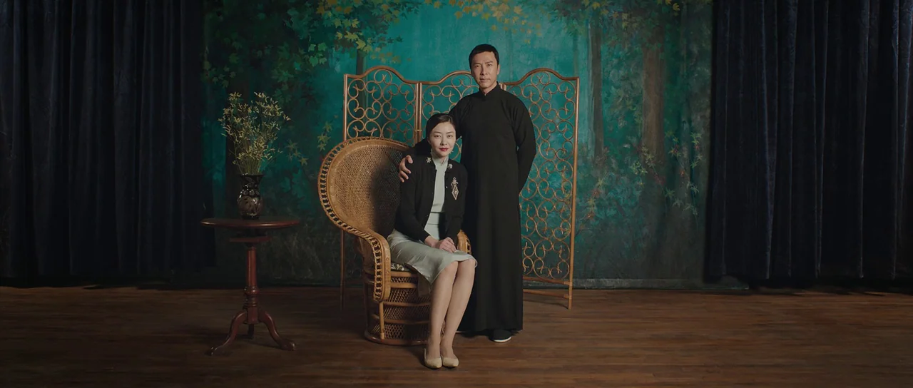 Ip Man 3 (2015) movie still: fullbody — A formal portrait studio features a hand-painted forest backdrop and a wicker divider. A man stands beside a…