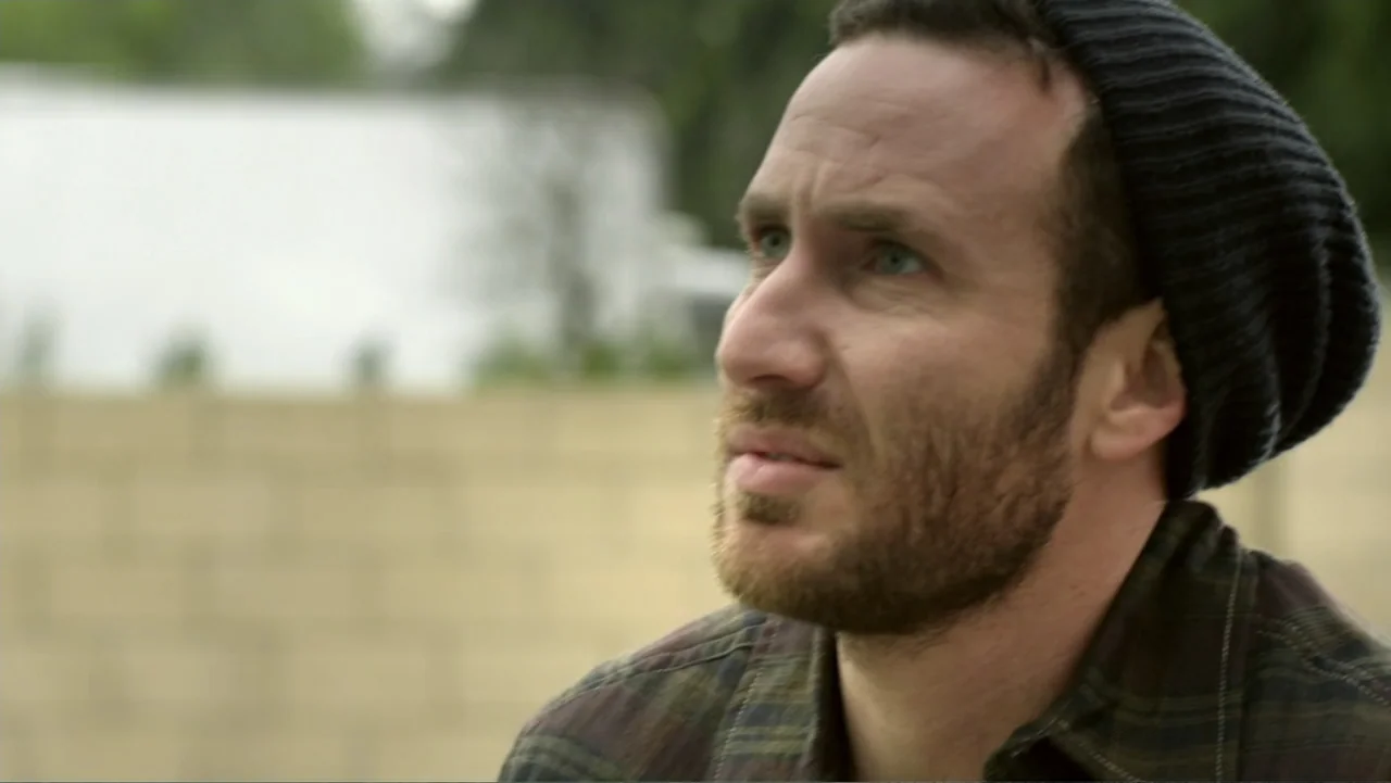 Free Samples (2013) movie still: closeup — An outdoor backyard setting features a blurred tan concrete block wall background. A man with a beard and…