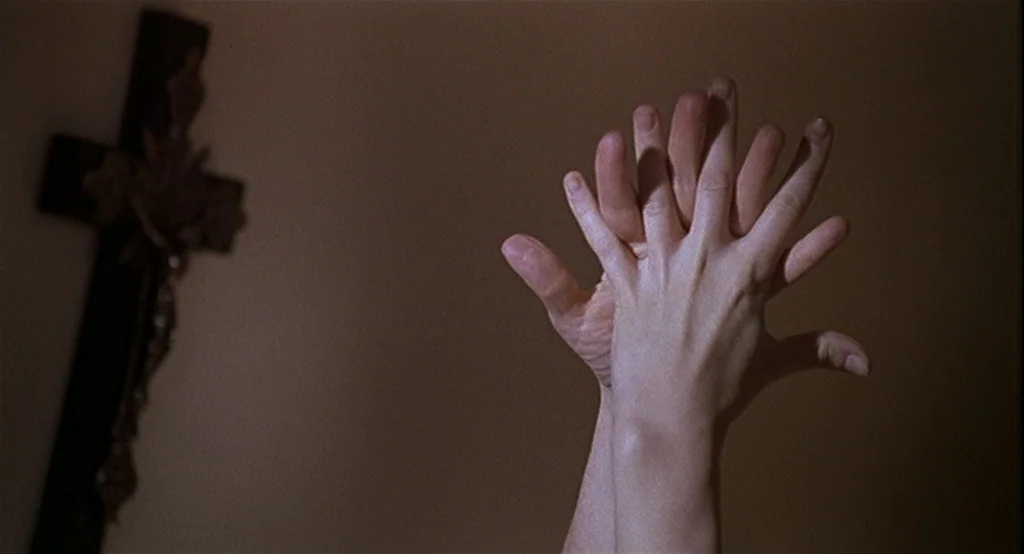 Deux (2002) movie still: closeup — A dimly lit room features a wooden wall cross hanging in the soft-focus background. Two pairs of hands press…