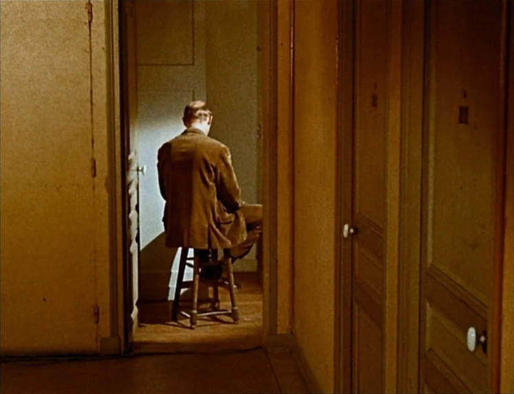 Zazie Dans Le Métro (1960) movie still: medium — A narrow domestic hallway features mustard-colored walls and multiple closed wooden doors. A man…