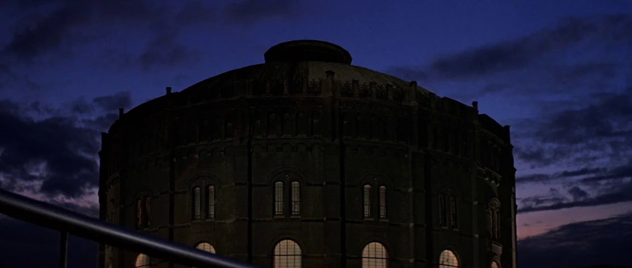 The Living Daylights (1987) movie still: establishing — A massive, cylindrical brick industrial building stands against a darkening purple evening…