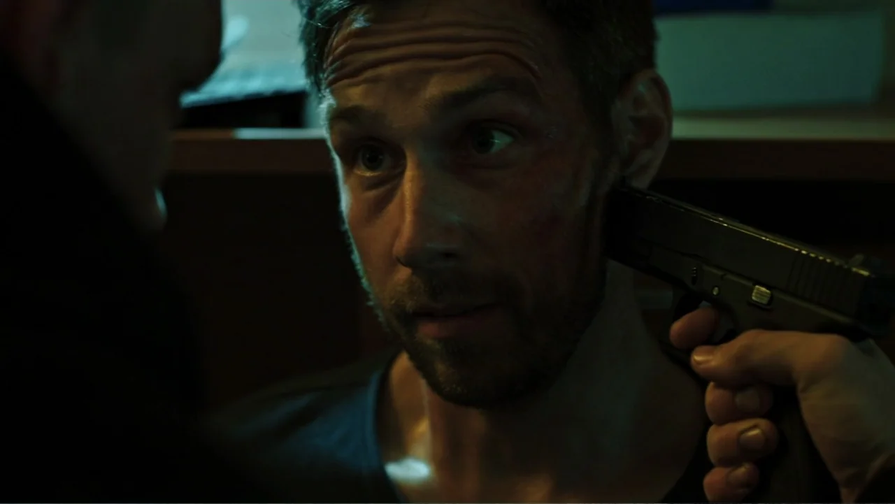 Bullet Proof (2022) movie still: closeup — A dimly lit room provides the backdrop for a tense confrontation between two men. A man is held at…