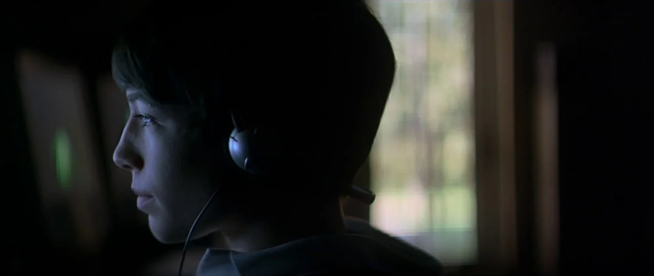 Afterschool (2008) movie still: closeup — A dark indoor space features a subject lit by the glow of a monitor. The person wears headphones while…
