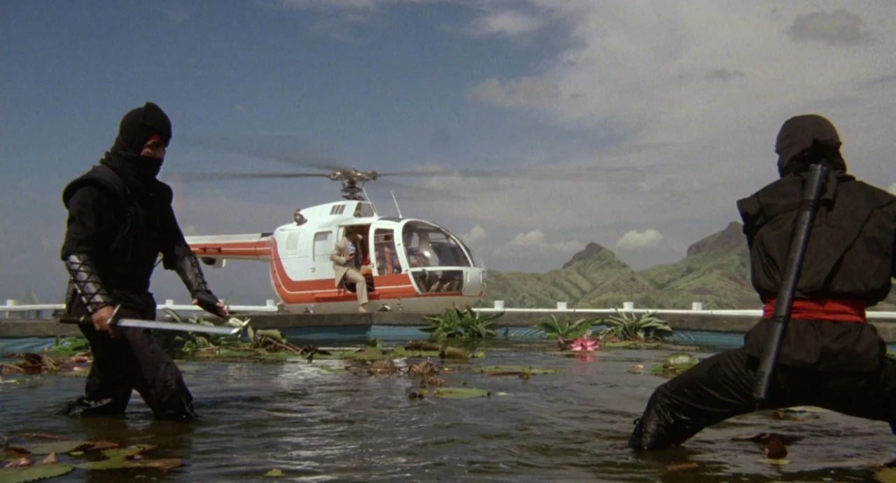 American Ninja (1985) movie still: wide — A shallow pond filled with lily pads sits under a clear blue sky. Two ninjas confront each other with…
