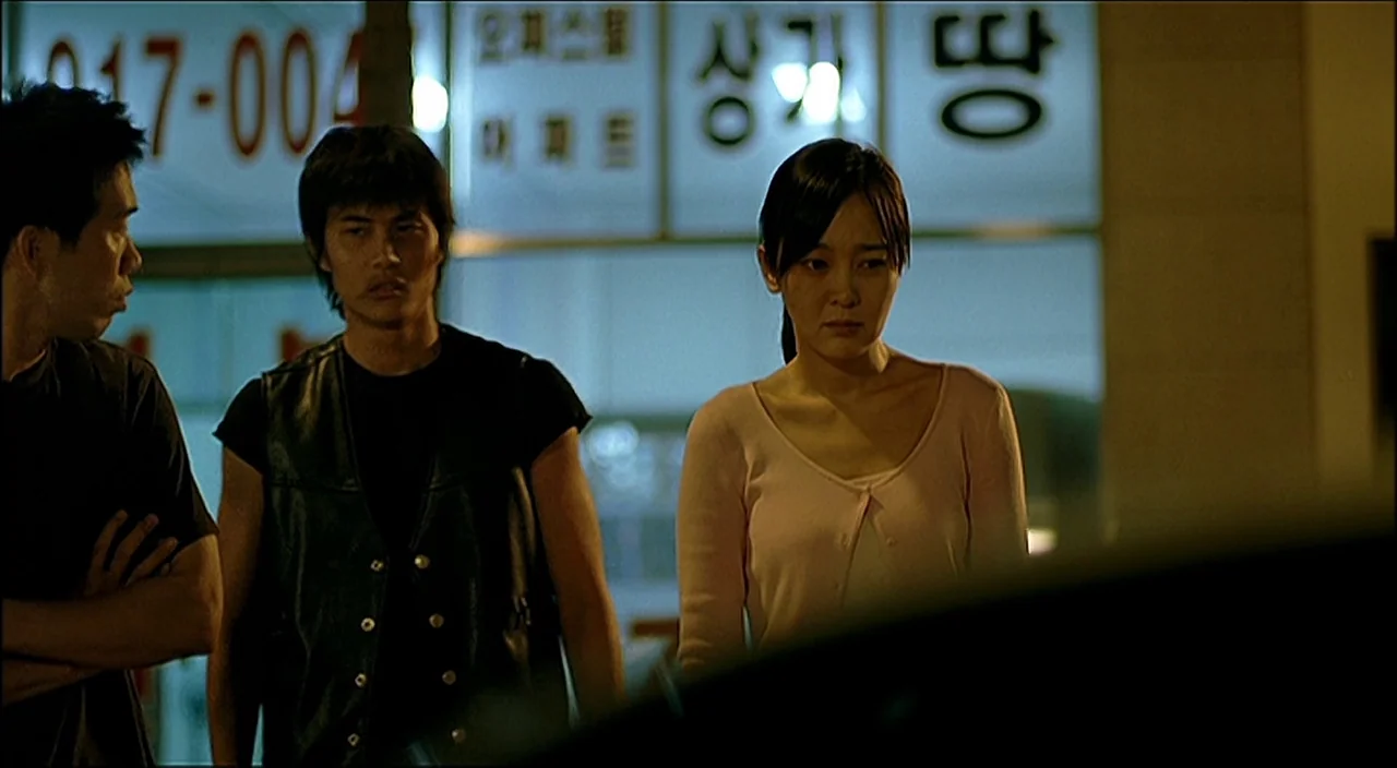 3-Iron (2004) movie still: medium — Three people stand in front of a storefront with white illuminated signs at night. A man in a leather vest…