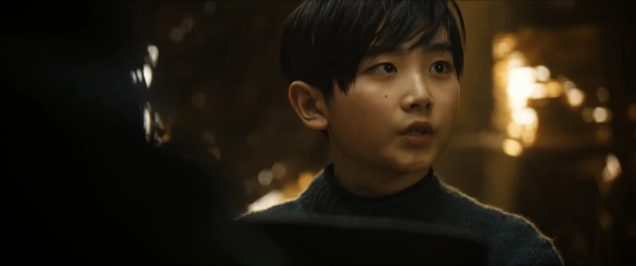 Star Trek: Section 31 (2025) movie still: closeup — A dark, dimly lit room with blurred warm lighting in the background. A young boy in a blue…