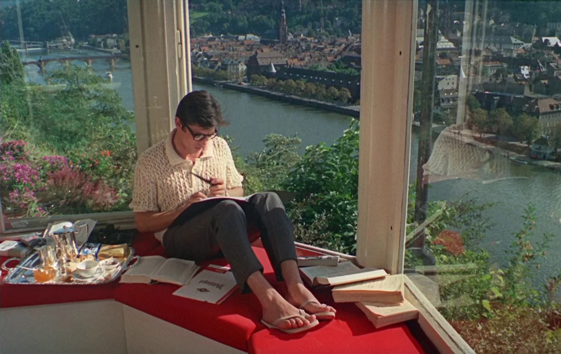 The Girl on a Motorcycle (1968) movie still: medium — A man sits on a red cushioned window bench overlooking a historic riverside city. Several…