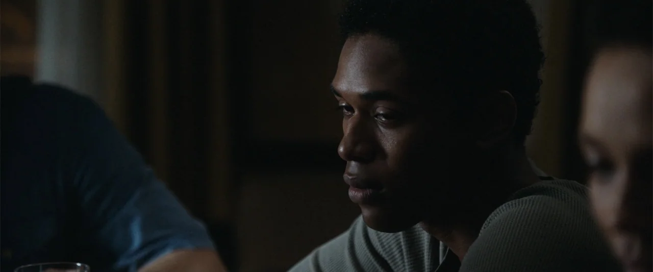 It Comes at Night (2017) movie still: closeup — A dark interior space with two people sitting near one another. A man in a tan shirt looks downward…