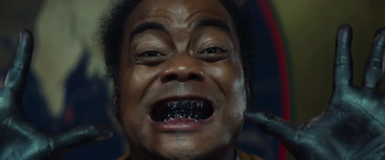 Kubi (2023) movie still: closeup — A man looks directly into the camera lens with his mouth held open wide. His teeth are stained black and his…