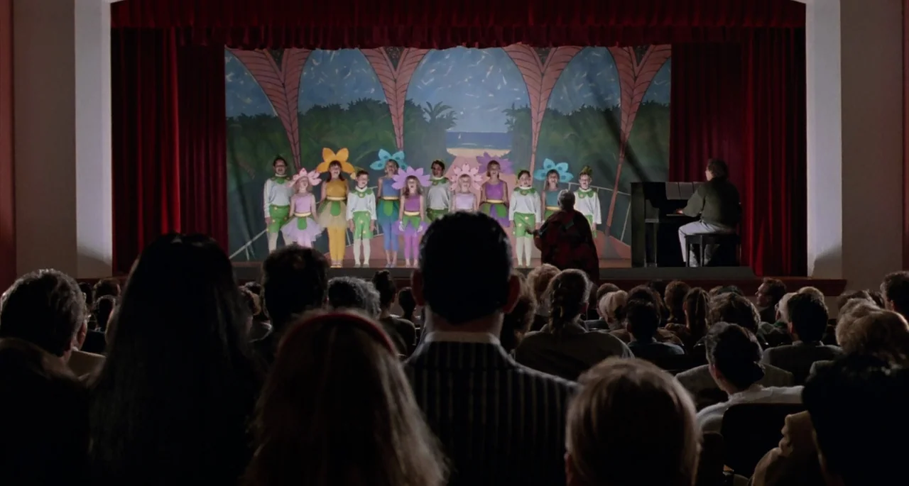The Addams Family (1991) movie still: wide — An auditorium features a stage with a painted backdrop showing a beach scene. A group of children…