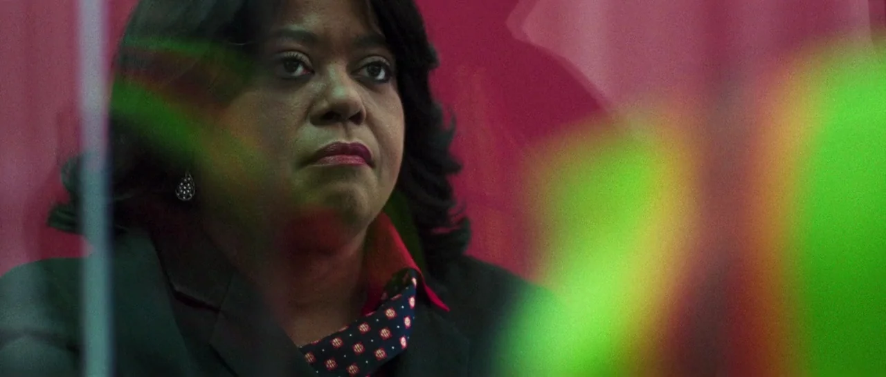 Good Time (2017) movie still: closeup — A woman sits in a dark, red-walled office space behind a glass barrier. She gazes forward while out-of-focus…