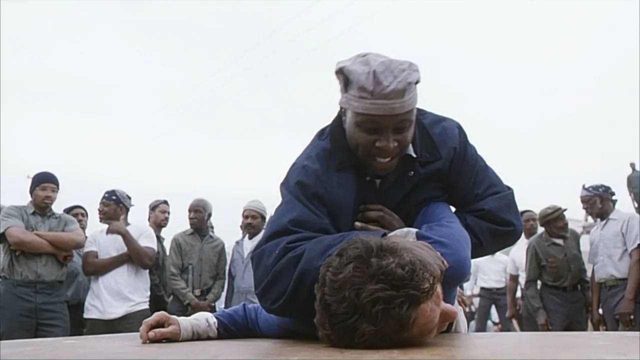 Against the Wall (1994) movie still: medium — An outdoor wooden platform is surrounded by a group of men watching. One man pins another face-down on…