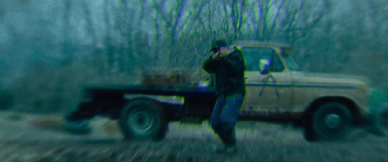 Dark Waters (2019) movie still: fullbody — A dark, blurred outdoor space contains a rusted flatbed truck parked in tall grass. The lone figure…