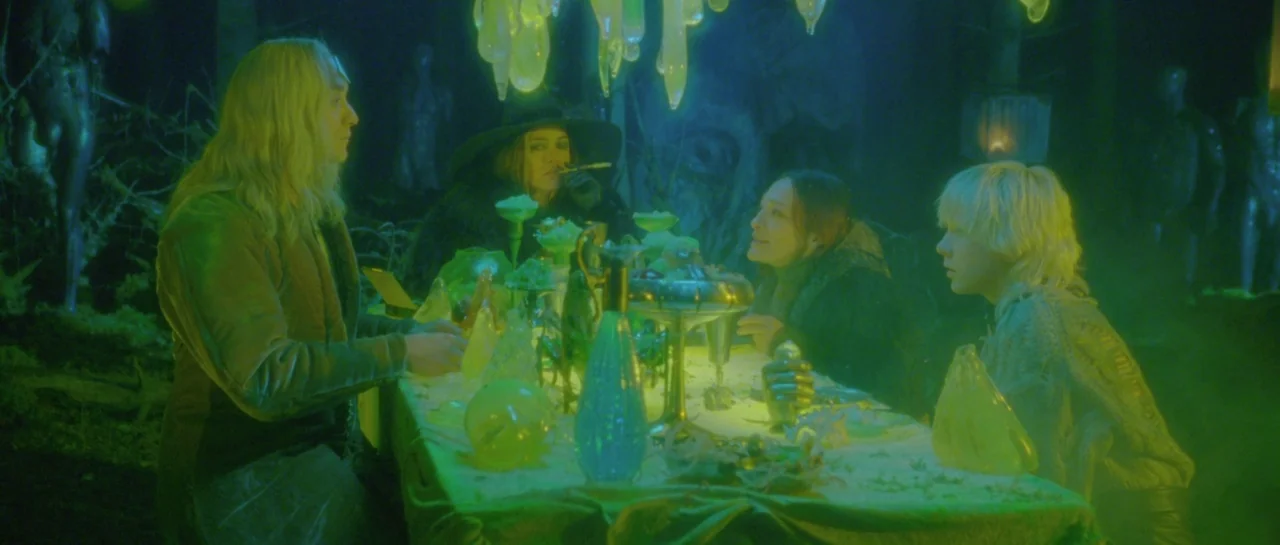 After Blue (2021) movie still: wide — Four people sit around a long covered table in a dark woodland environment. Strange glowing translucent…