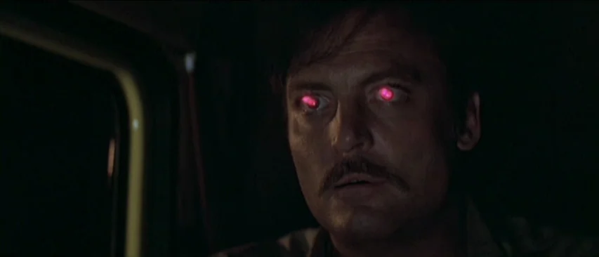 Roadgames (1981) movie still: closeup — The interior of a vehicle is shrouded in near total darkness. A man with glowing red eyes stares forward in…