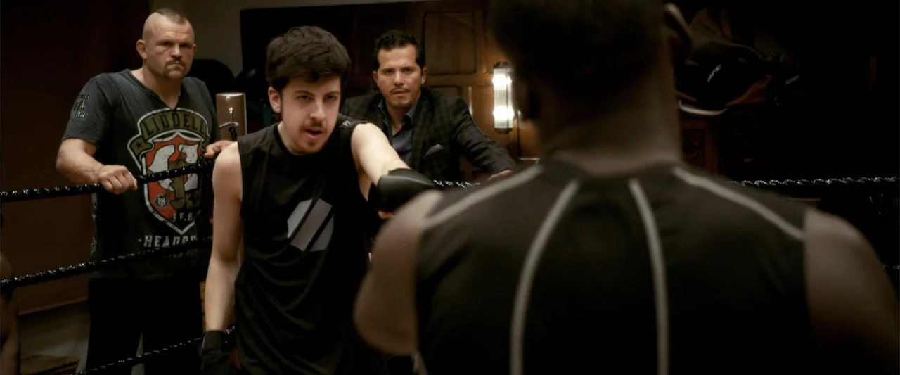 Kick-Ass 2 (2013) movie still: medium — A dimly lit indoor space features a rope-enclosed fighting area with several men present. A young man…