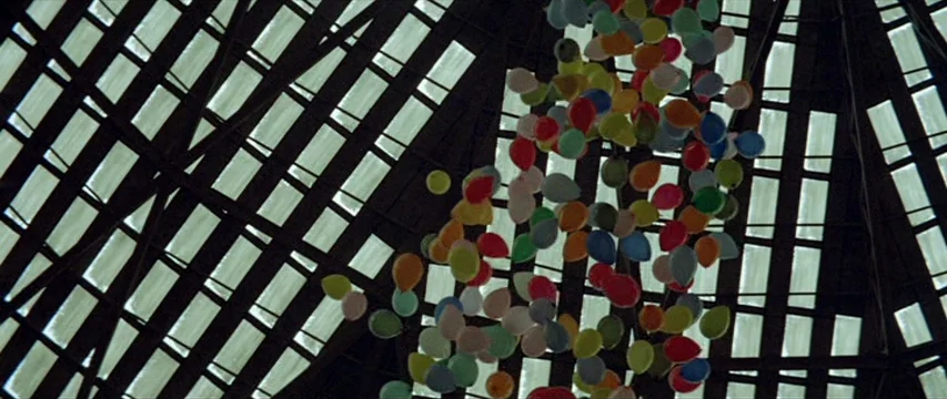 Brewster McCloud (1970) movie still: establishing — A high, grid-patterned architectural ceiling frames a dense cluster of multi-colored balloons.…