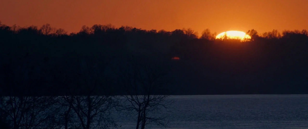Harriet movie still: establishing — A dark treeline sits along the edge of a calm, dark water surface. The sun glows intensely orange as it dips…