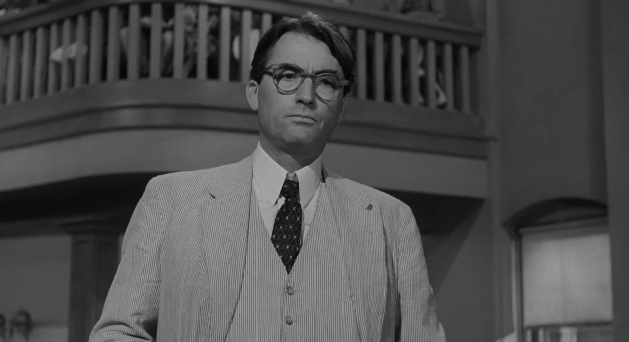 To Kill A Mockingbird (1962) movie still: medium — An interior space with a wooden balcony railing visible behind a man. The man stands in the…