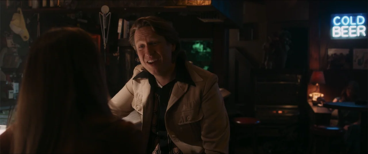 Woman of the Hour (2024) movie still: medium — A dark dive bar interior features wooden surfaces and a glowing blue neon sign. A man in a tan jacket…