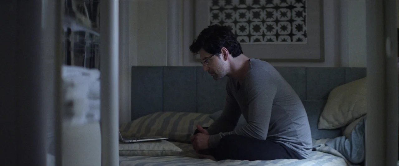 Chevalier (2015) movie still: medium — A man sits on a bed in a dimly lit, contemporary bedroom. He is hunched over and looking down at a laptop…