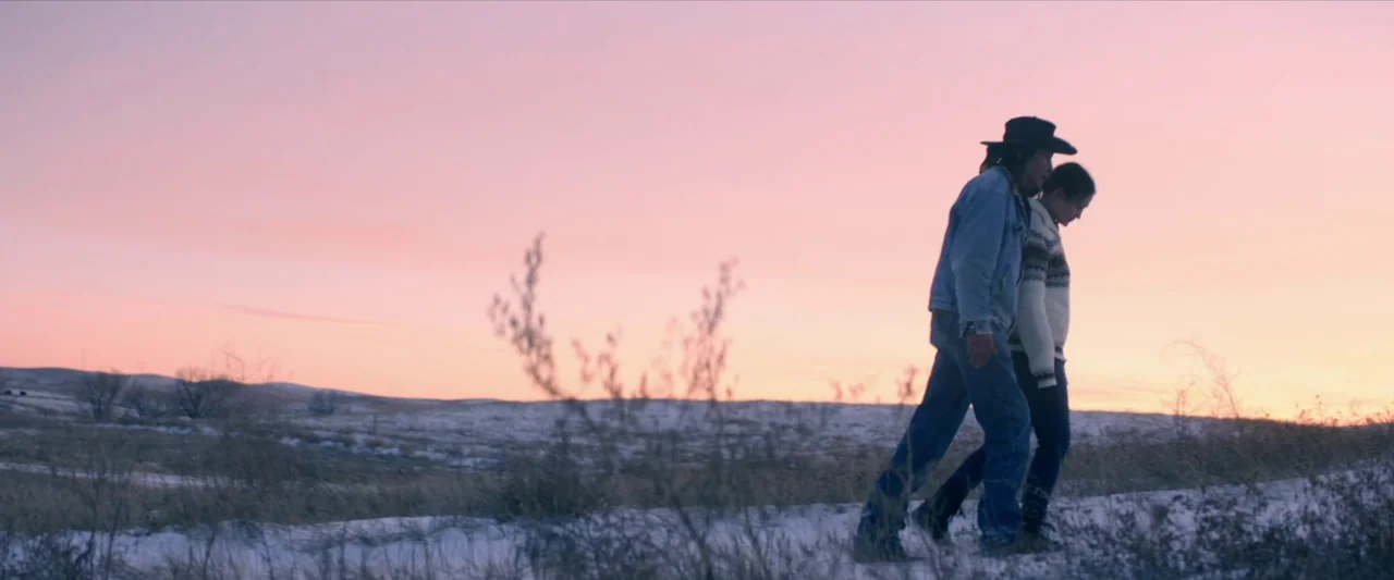 The Unknown Country (2022) movie still: fullbody — A snow-dusted, grassy landscape under a vibrant pink and orange sunset sky. A man in a cowboy hat…