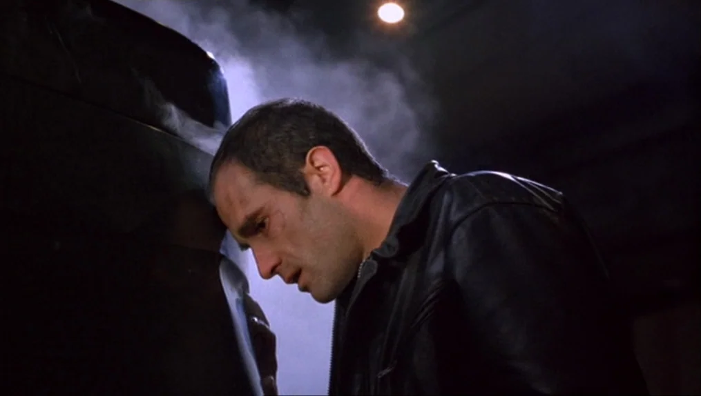 Crash (1996) movie still: medium — A dark, dimly lit parking structure at night with thin wisps of smoke present. A man in a leather jacket presses…