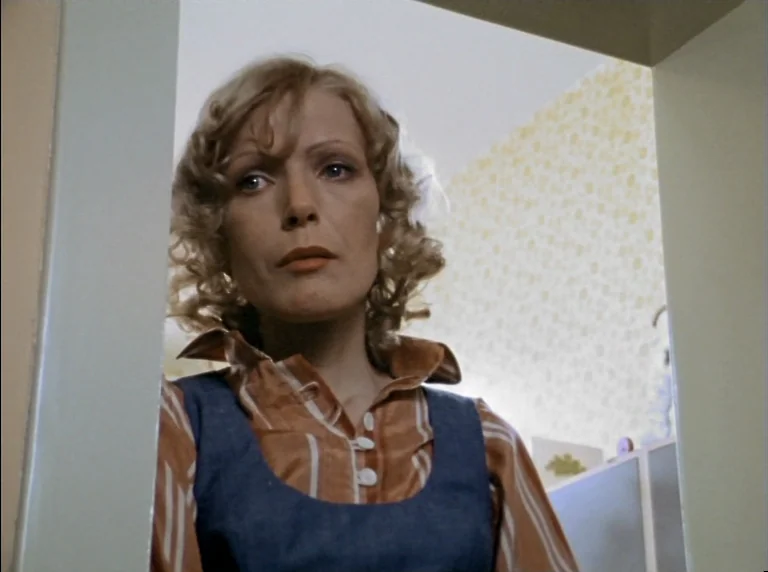 Fear Of Fear (1975) movie still: medium — A woman stands in a doorway looking into a room with patterned wallpaper. She is wearing a striped…