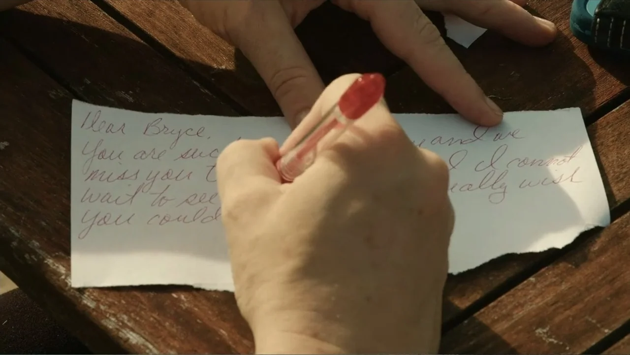 Death of a Child (2017) movie still: closeup — A weathered wooden outdoor table surface provides the backdrop for a piece of paper. A person's hand…