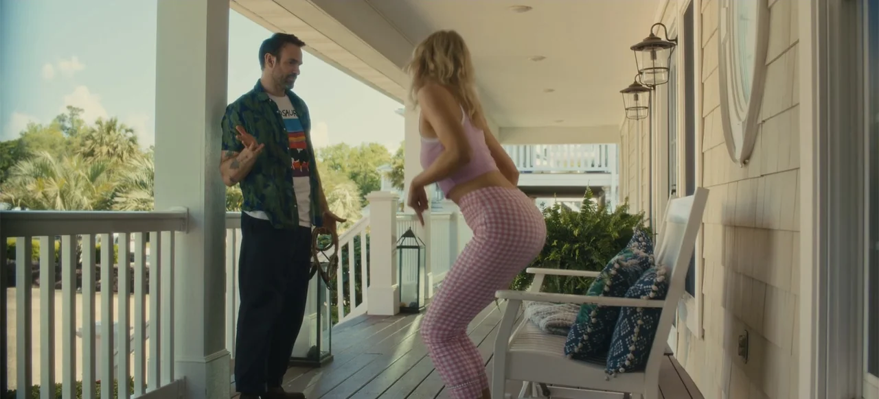 Merv (2025) movie still: fullbody — A wooden porch with a white railing and a white rocking bench sits outdoors. A young woman in patterned leggings…