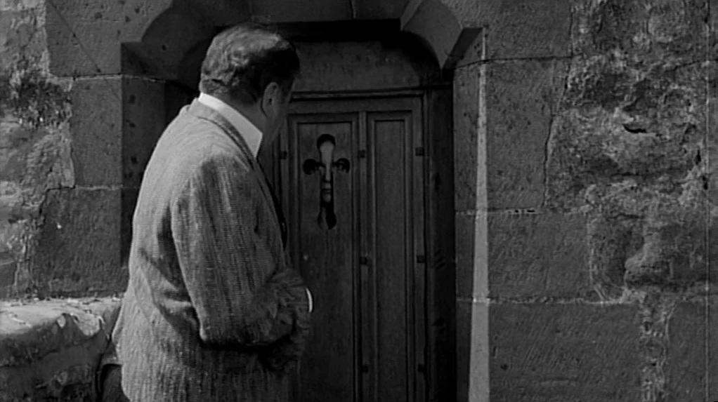 Cul-de-sac (1966) movie still: medium — A man stands before a stone structure with a wooden door featuring a cutout. The cutout shaped like a cross…