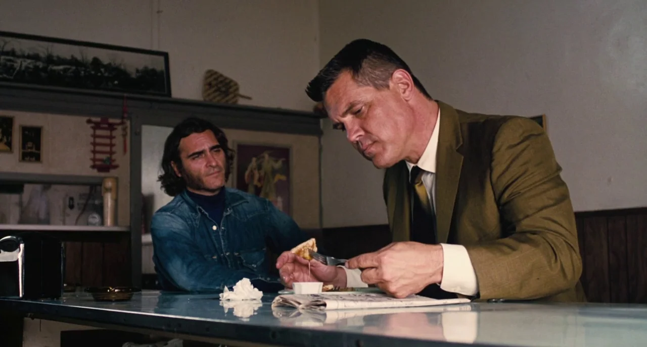 Inherent Vice (2014) movie still: medium — A dimly lit diner with wood-paneled walls and a worn counter. A man in a suit eats while another man…