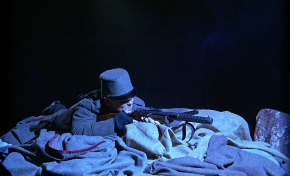 Wittgenstein (1993) movie still: medium — A soldier positions himself behind a row of sandbags in a dark, nocturnal environment. He grips a…