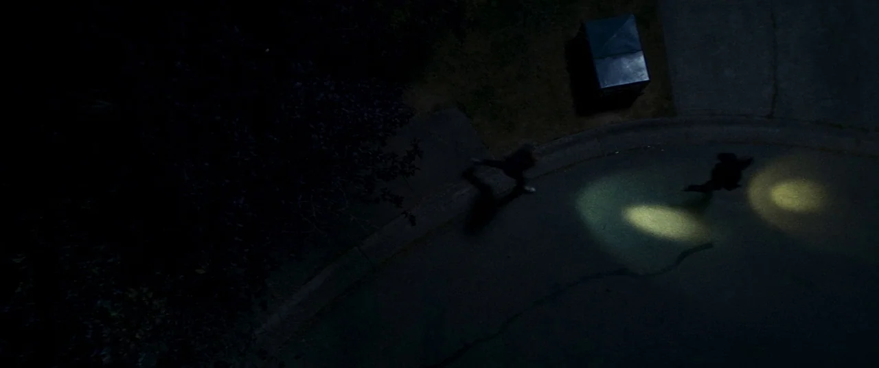 Summer of 84 (2018) movie still: wide — A nighttime suburban cul-de-sac features a dark asphalt road and a nearby trash bin. Two silhouetted…