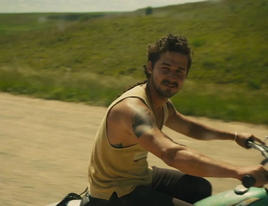 American Honey (2016) movie still: medium — A man rides a motorbike along a dusty dirt track through a rural landscape. The rolling green hills…