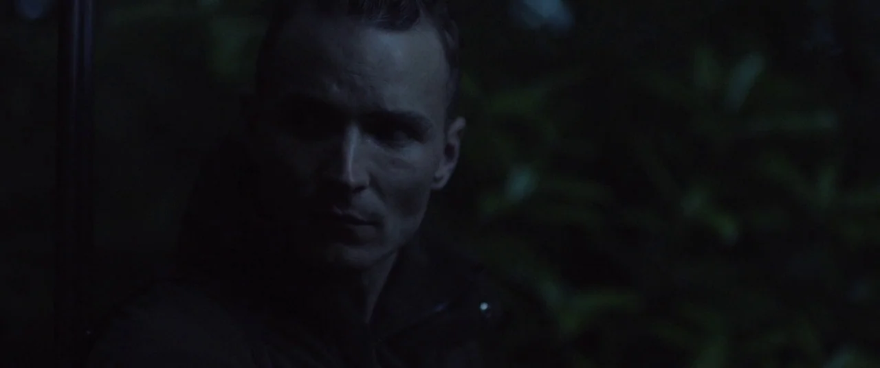 The Survivalist (2015) movie still: closeup — A man stands in dense, dark vegetation at night under low light. He looks away from the frame while…
