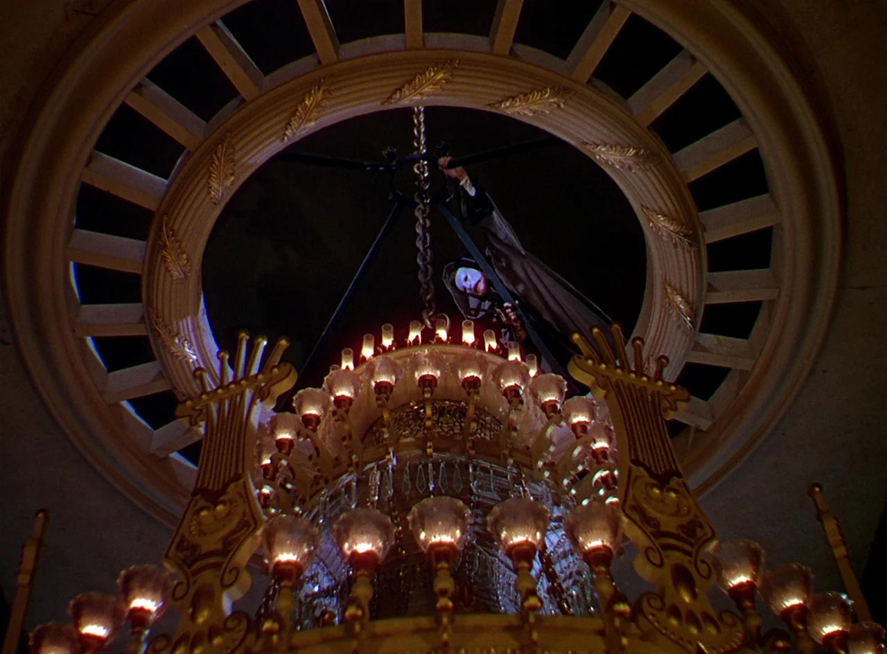 Phantom of the Opera (1943) movie still: wide — A man wearing a white mask clings to a chain above an ornate chandelier. The chandelier is…