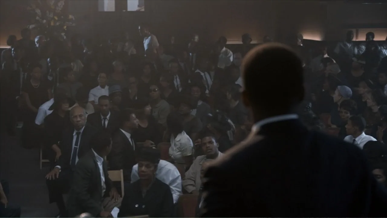 All the Way (2016) movie still: medium — A dark interior filled with rows of people in formal attire seated on wooden chairs. A man in a suit stands…