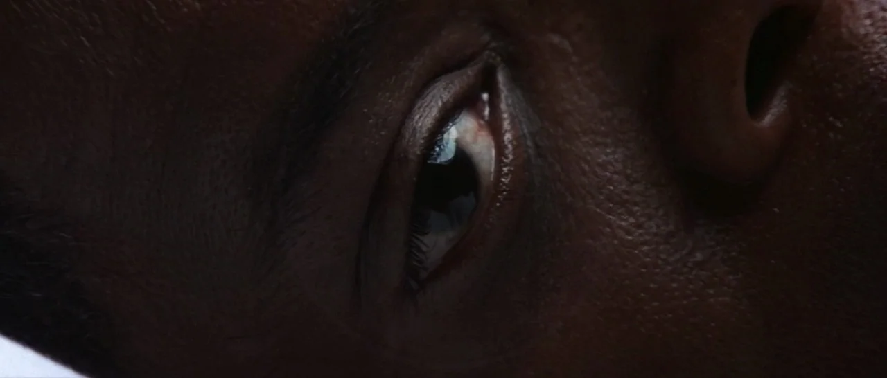 Unbreakable (2000) movie still: closeup — An extreme close-up focuses on a person's eye in dark, dimly lit conditions. The surrounding skin and…