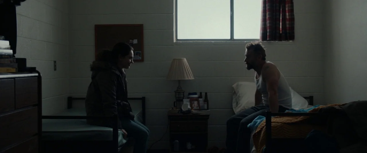 Little Woods (2018) movie still: wide — A spartan motel room with white cinderblock walls and two simple single beds. The man and woman sit facing…