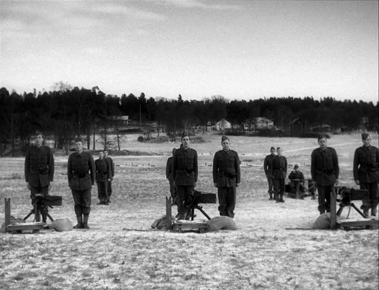 Music in Darkness (1948) movie still: wide — A group of soldiers stands in formation on a vast, snow-dusted, sparse field. Each man stands behind a…