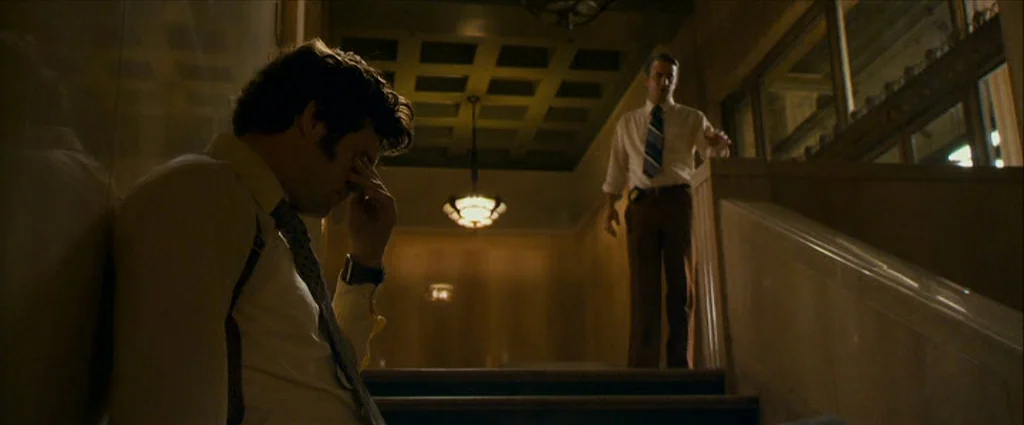 Zodiac (2007) movie still: medium — A dimly lit municipal stairwell with stone walls and a coffered ceiling. One man sits on the steps hiding his…