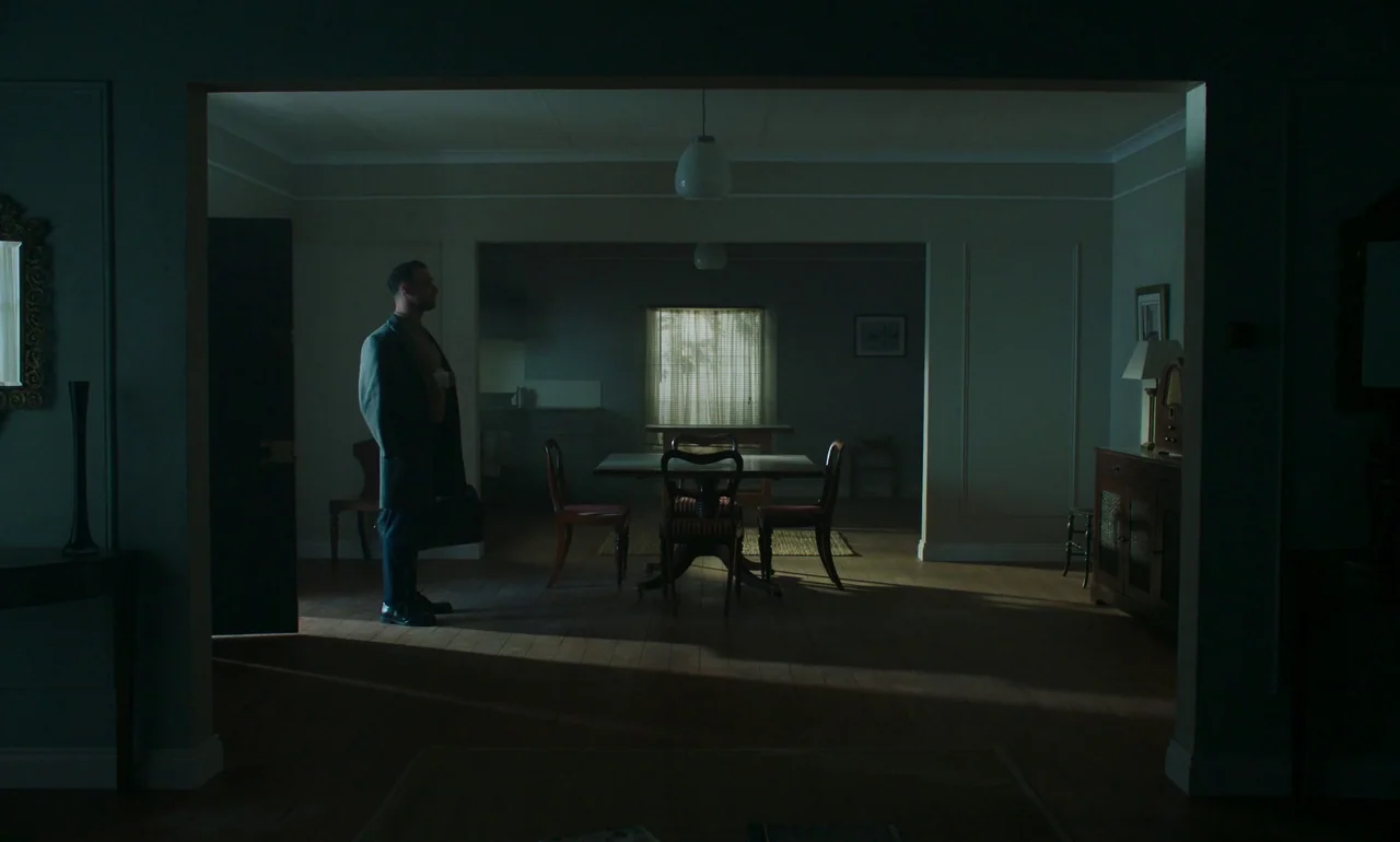 It Is In Us All (2022) movie still: wide — A spacious, sparsely furnished dining area features wooden floors and dark, muted teal walls. A lone man…