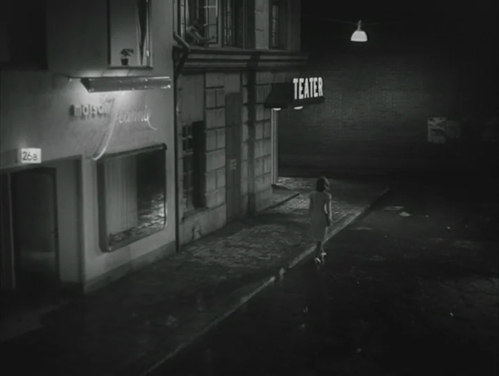 Crisis (1946) movie still: wide — An empty nighttime city street features a theater sign and a boutique storefront. A woman walks alone away from…