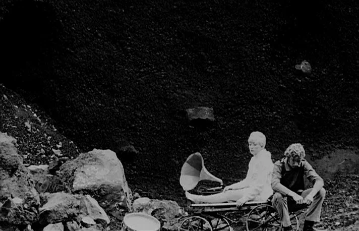Fando y Lis (1968) movie still: wide — A dark, rocky industrial quarry landscape dominates the background with large stone boulders present. A woman…