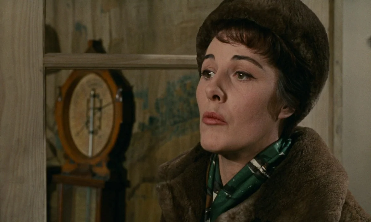Muriel (1963) movie still: closeup — A woman stands indoors in front of a wooden wall partition. A vintage round wooden wall clock is visible in the…