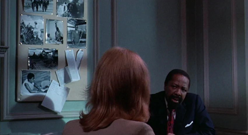 The Passenger (1975) movie still: medium — An interior office space with a paneled green wall and a bulletin board covered in photographs. A man…