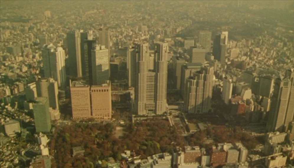 Gozu (2003) movie still: establishing — A high-altitude view captures a sprawling metropolitan city dominated by large skyscraper structures. The…