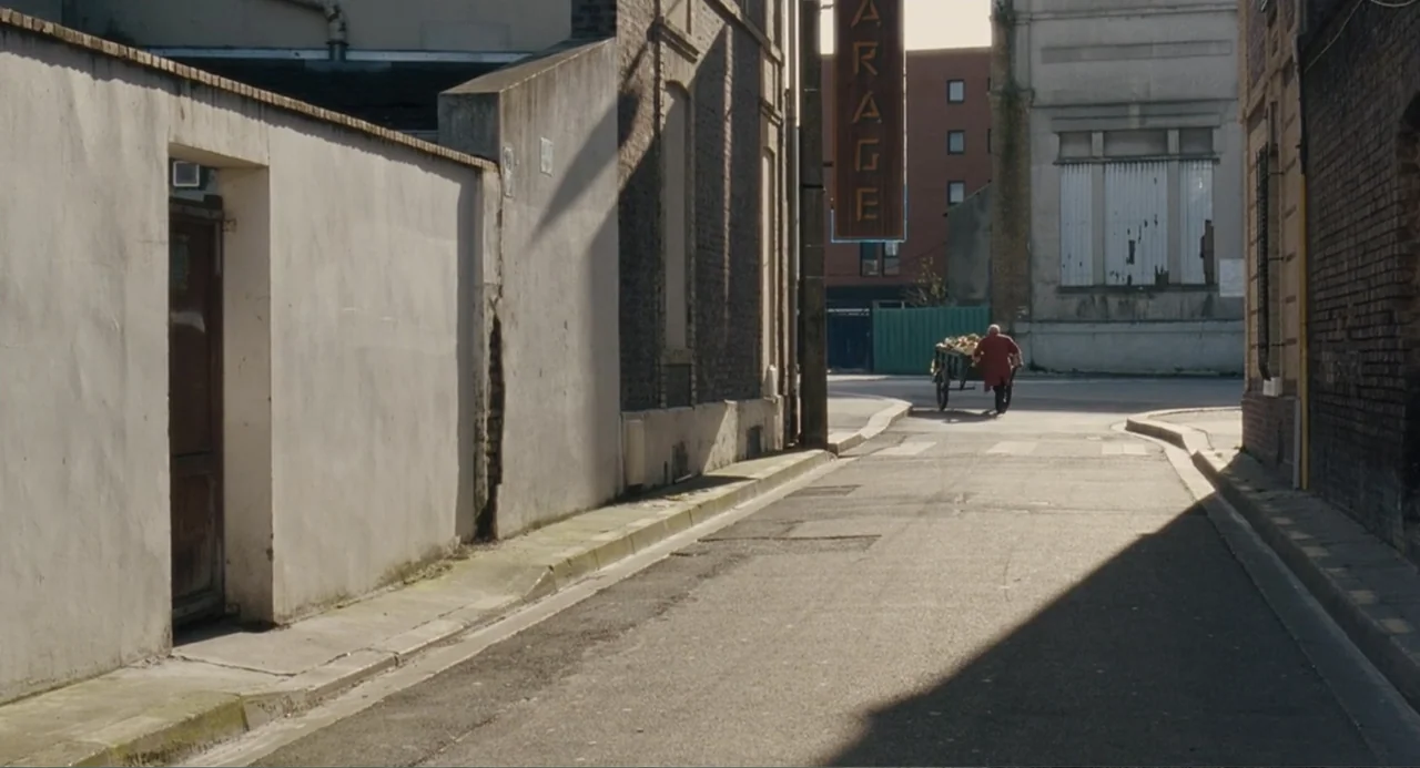 Le Havre (2011) movie still: wide — A narrow paved street features a tall white wall and brick buildings under sunlight. An elderly man walks away…